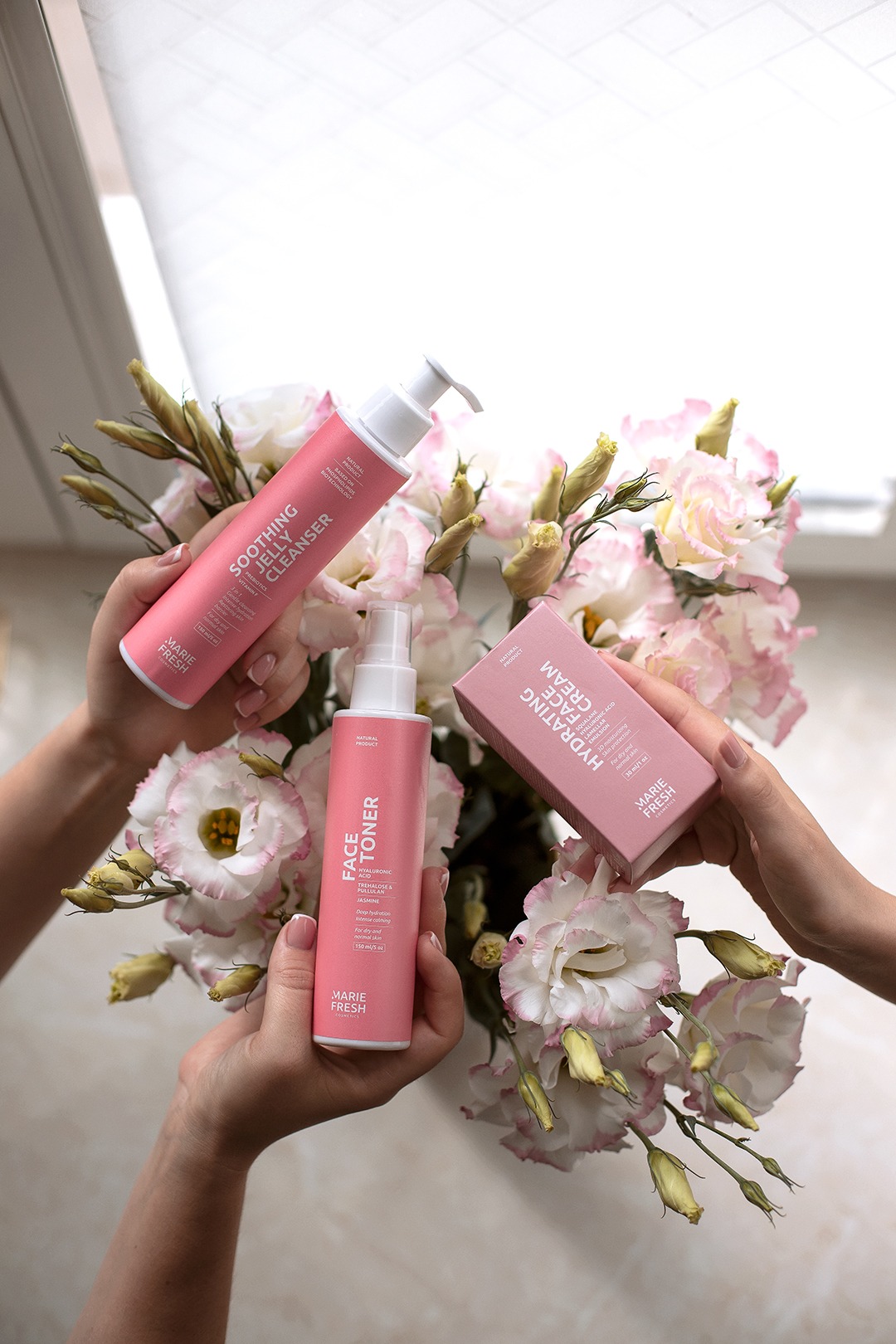 Dłonie prezentują zestaw kosmetyków Marie Fresh: żel oczyszczający, tonik do twarzy i krem nawilżający na tle bukietu białych róż z różowymi brzegami płatków.