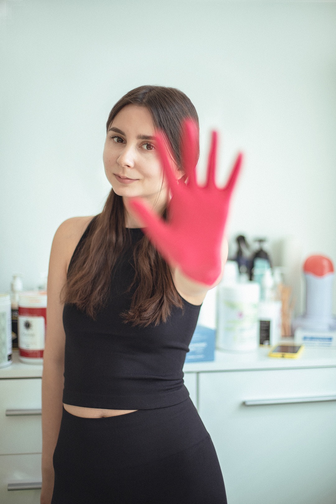 Kobieta w czarnym stroju sportowym z wyciągniętą dłonią w różowej rękawiczce na tle białej szafki z kosmetykami i telefonem.