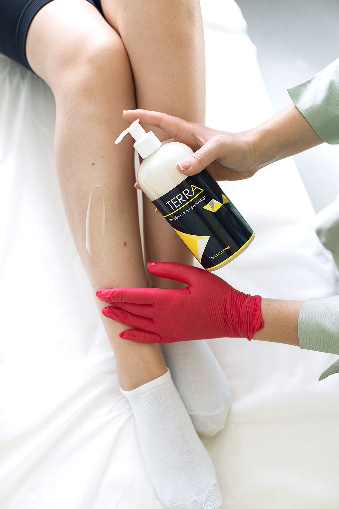 Nakładanie kremu po depilacji na nogę przez osobę w czerwonej rękawiczce, widoczna butelka kremu marki Terra
