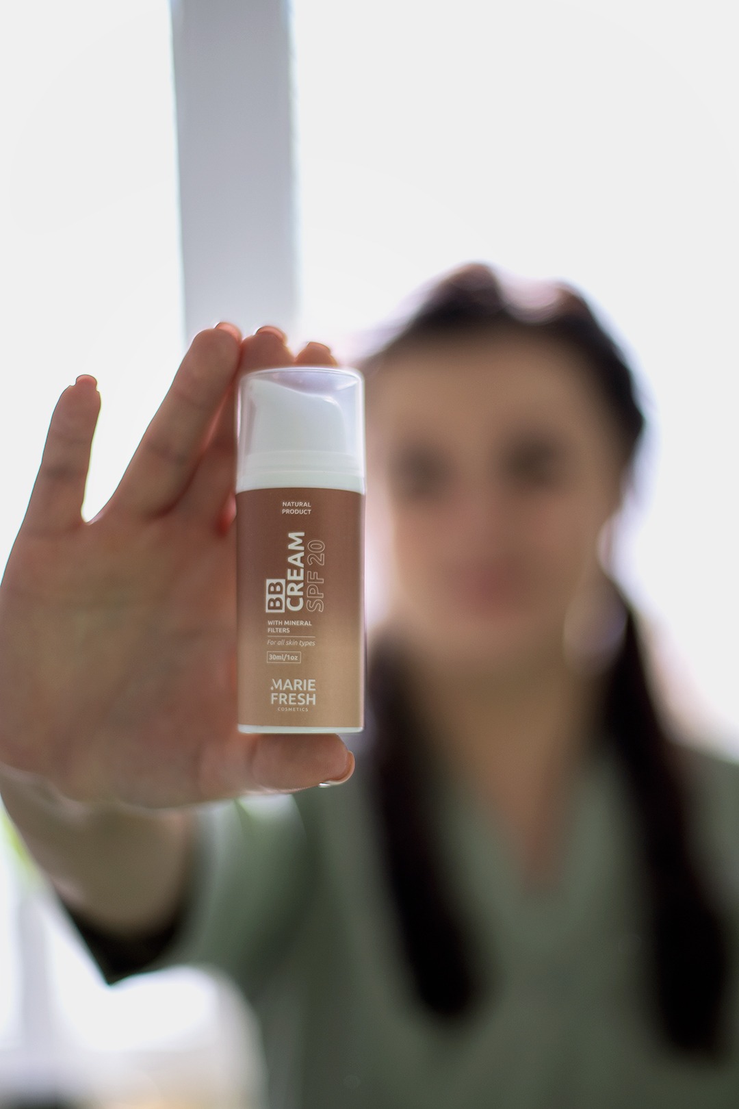 Dłoń prezentuje tubkę beżowego kremu BB z filtrem SPF 20 marki Marie Fresh Cosmetics na rozmytym tle twarzy kobiety i jasnego okna.