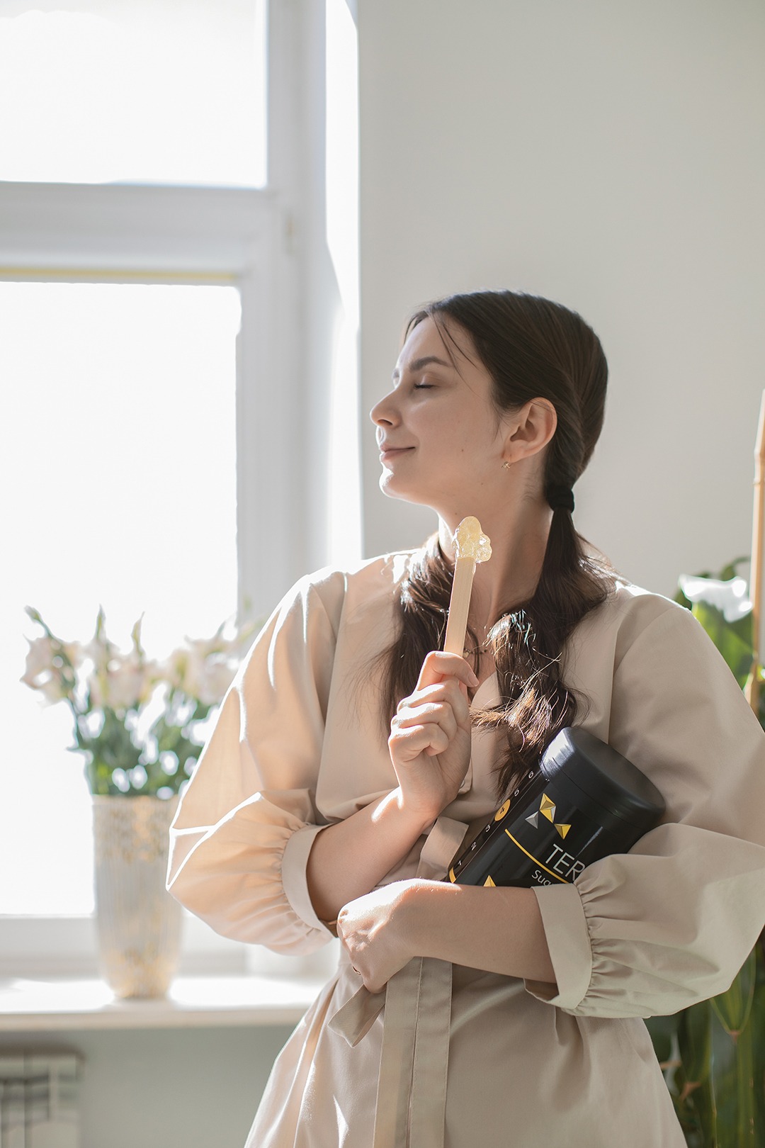 Kobieta z zamkniętymi oczami trzyma szpatułkę z woskiem i pojemnik z woskiem do depilacji w jasnym wnętrzu.