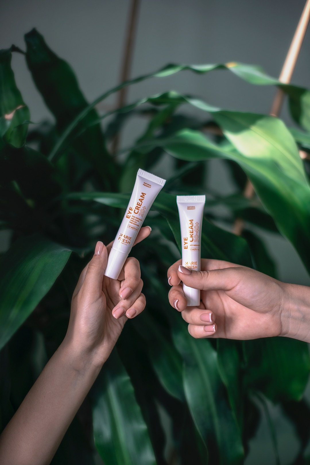 Dwie dłonie trzymają tubki kremu pod oczy marki Marie Fresh na tle zielonych liści rośliny doniczkowej. Jedna tubka oznaczona 20+, druga 30-40+.