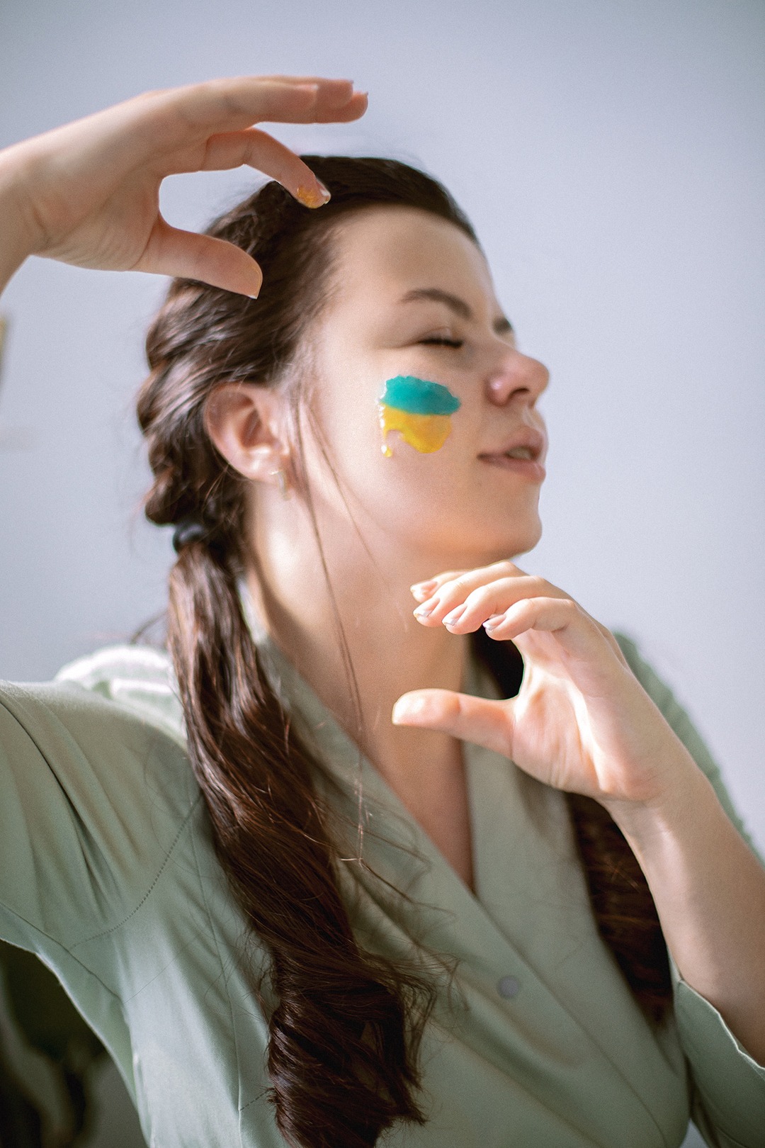 Portret kobiety z zamkniętymi oczami i flagą Ukrainy namalowaną na policzku, dłonie ułożone w ramkę wokół twarzy, włosy splecione w warkocze.