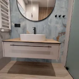 Nowoczesna łazienka w Lesku z umywalką nablatową na szafce z jasnego drewna i białym frontem, okrągłe lustro z czarną ramą i dekoracyjne płytki imitujące marmur na ścianie.