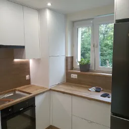 Nowoczesna, narożna kuchnia z białymi frontami, drewnianym blatem i brązowymi panelami ściennymi, z widocznym piekarnikiem, oświetleniem LED pod szafkami i rośliną w doniczce na parapecie.