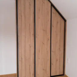 Trzydrzwiowa szafa przesuwna z drewnopodobnymi frontami i czarnymi obramowaniami, dopasowana do skosu poddasza, widoczna z perspektywy podłogi wyłożonej panelami.