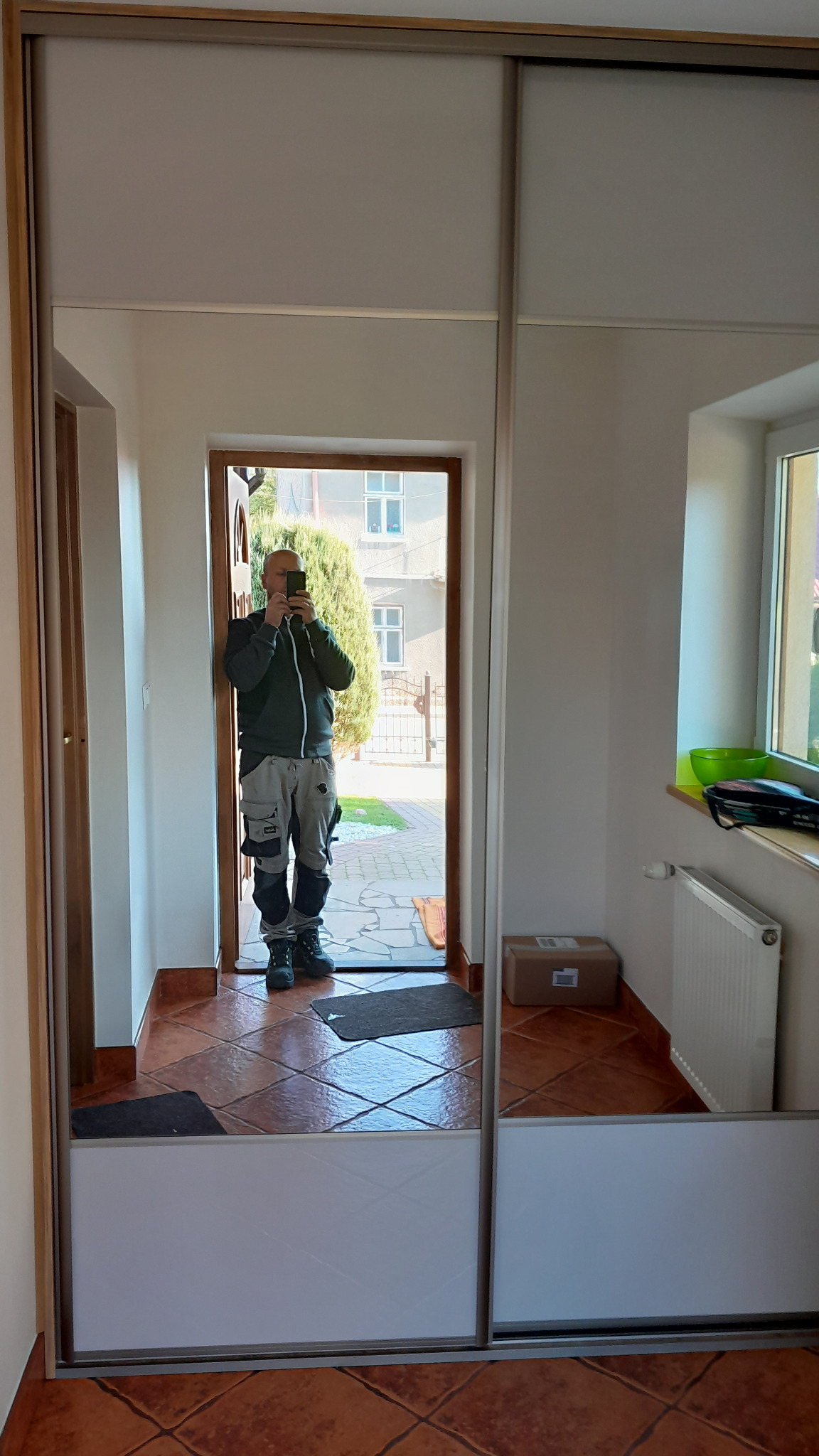 Mężczyzna w odzieży roboczej robi zdjęcie smartfonem w lustrzanych drzwiach szafy przesuwnej, widoczne płytki podłogowe w odcieniach brązu i fragment przedpokoju z oknem.