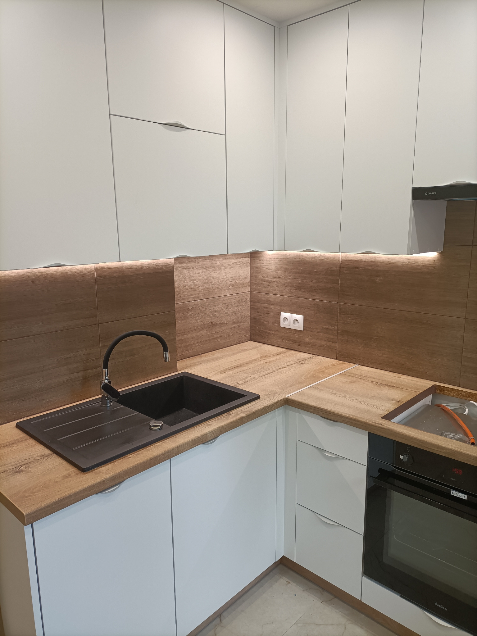 Nowoczesna zabudowa kuchenna z białymi szafkami z subtelnymi uchwytami, drewnianym blatem i brązowymi panelami ściennymi z podświetleniem LED, czarnym zlewem i piekarnikiem Amica.