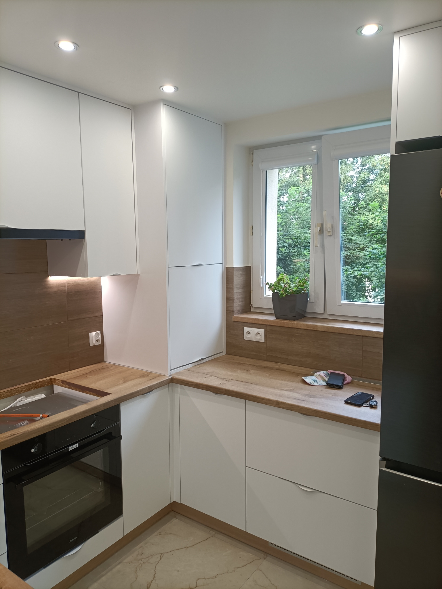 Nowoczesna, narożna kuchnia z białymi frontami, drewnianym blatem i brązowymi panelami ściennymi, z widocznym piekarnikiem, oświetleniem LED pod szafkami i rośliną w doniczce na parapecie.