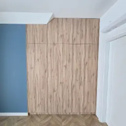 Wysoka szafa na wymiar z drewnopodobnymi frontami, dopasowana do skosu sufitu, zintegrowana z niebieską ścianą i parkietem ułożonym w jodełkę.