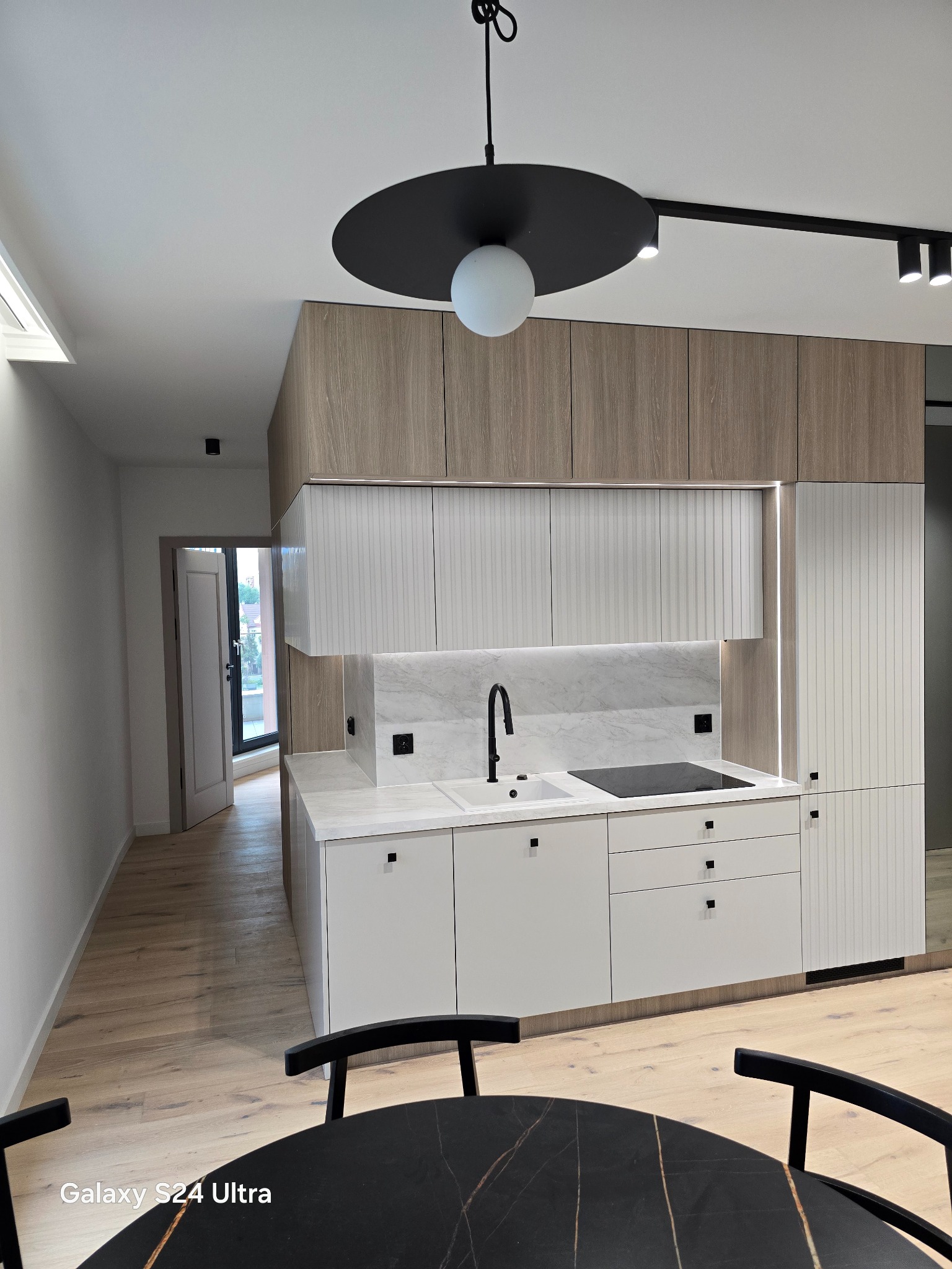 Nowoczesna kuchnia z białymi szafkami i drewnianymi górnymi panelami, czarna bateria i płyta indukcyjna na marmurowym blacie, oświetlenie podszafkowe i czarny okrągły stół z krzesłami w tle.