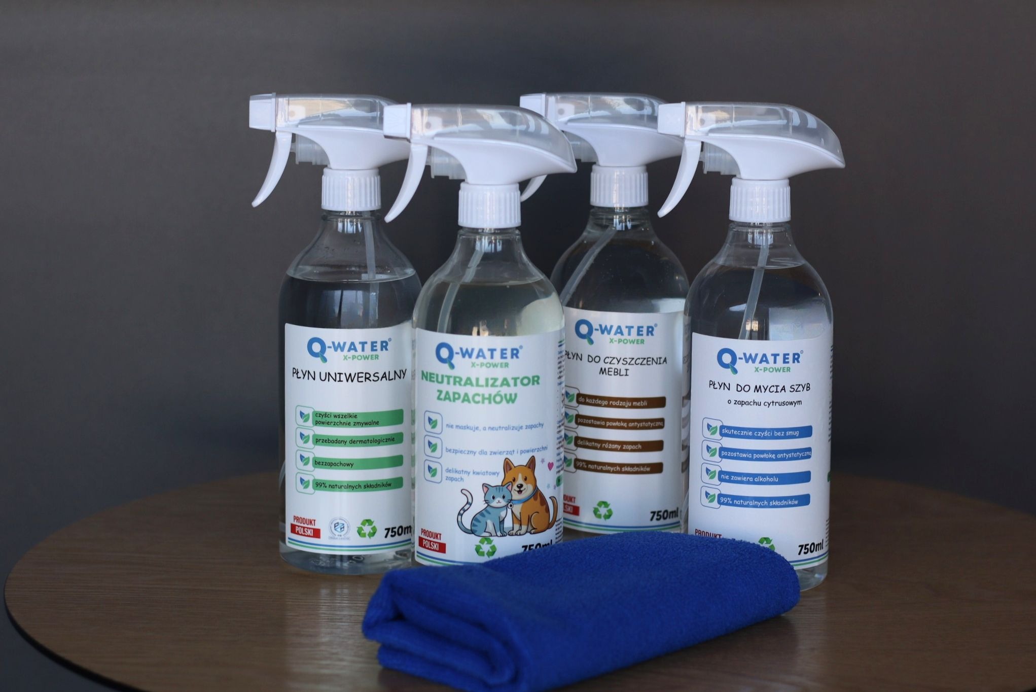 Cztery butelki z rozpylaczem Q-Water: uniwersalny, neutralizator zapachów, do mebli i szyb. Niebieska ściereczka z mikrofibry na drewnianym blacie.