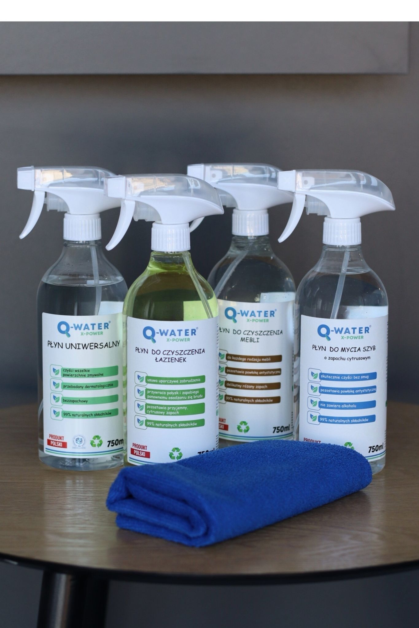 Zestaw czterech butelek Q-Water z rozpylaczem: płyn uniwersalny, do łazienek, mebli i szyb. Niebieska ściereczka z mikrofibry na drewnianym stole.