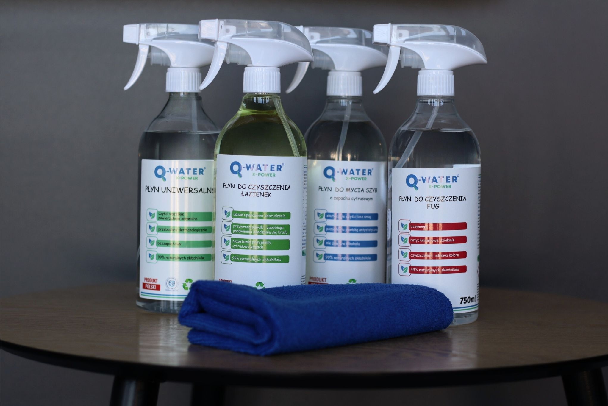 Cztery butelki z rozpylaczem Q-Water X-Power: płyn uniwersalny, do łazienek, do szyb i do fug. Obok leży złożona, niebieska ściereczka z mikrofibry.