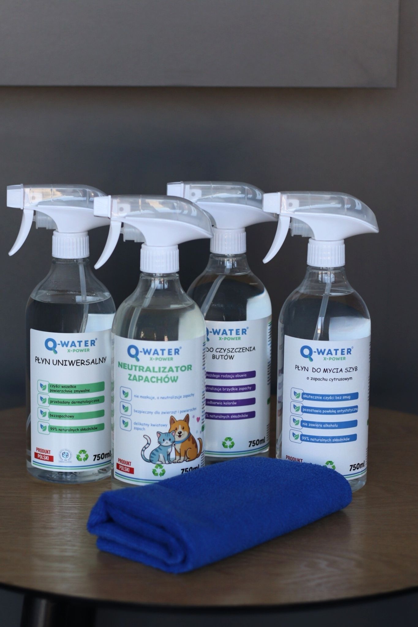 Cztery butelki Q-Water z rozpylaczem: uniwersalny, neutralizator zapachów, do butów i szyb. Niebieska ściereczka z mikrofibry na drewnianym stole.