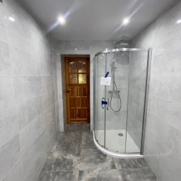 MICHAŁ SKÓRA TransBUD - Nowoczesna łazienka z przeszkloną kabiną prysznicową, wykończona szarymi płytkami na ścianach i podłodze, oświetlona punktowymi lampami sufitowymi, z widocznymi detalami drewnianych drzwi w tle.