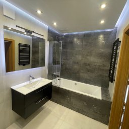 MICHAŁ SKÓRA TransBUD - Nowoczesna łazienka z szarymi płytkami imitującymi kamień, białą wanną z parawanem, czarną szafką pod umywalką i dużym lustrem z oświetleniem LED.