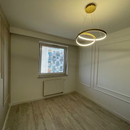MICHAŁ SKÓRA TransBUD - Pusty pokój z panelami ściennymi w stylu klasycznym, parkietem w odcieniach beżu i nowoczesną lampą sufitową z dwoma okręgami LED.