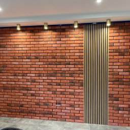 MICHAŁ SKÓRA TransBUD - Ściana z cegły klinkierowej z pionowym panelem dekoracyjnym z drewnianych listew, oświetlona punktowymi lampami sufitowymi.