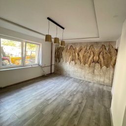 MICHAŁ SKÓRA TransBUD - Pusty pokój po remoncie z panelami podłogowymi imitującymi drewno, tapetą w złote liście na jednej ścianie i trzema złotymi lampami wiszącymi.