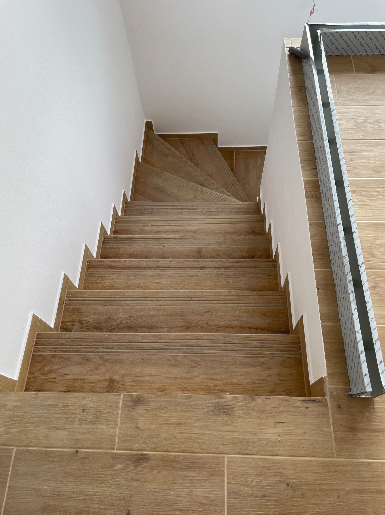 Schody wykończone płytkami imitującymi drewno, z białymi ścianami i listwami przypodłogowymi, widok z góry, z elementami zabezpieczającymi na podłodze.