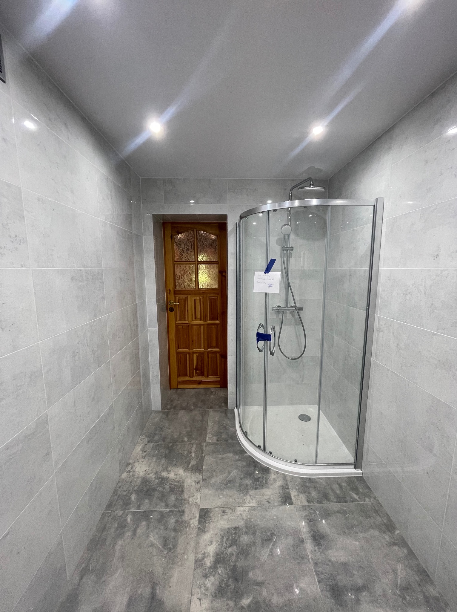 Nowoczesna łazienka z przeszkloną kabiną prysznicową, wykończona szarymi płytkami na ścianach i podłodze, oświetlona punktowymi lampami sufitowymi, z widocznymi detalami drewnianych drzwi w tle.