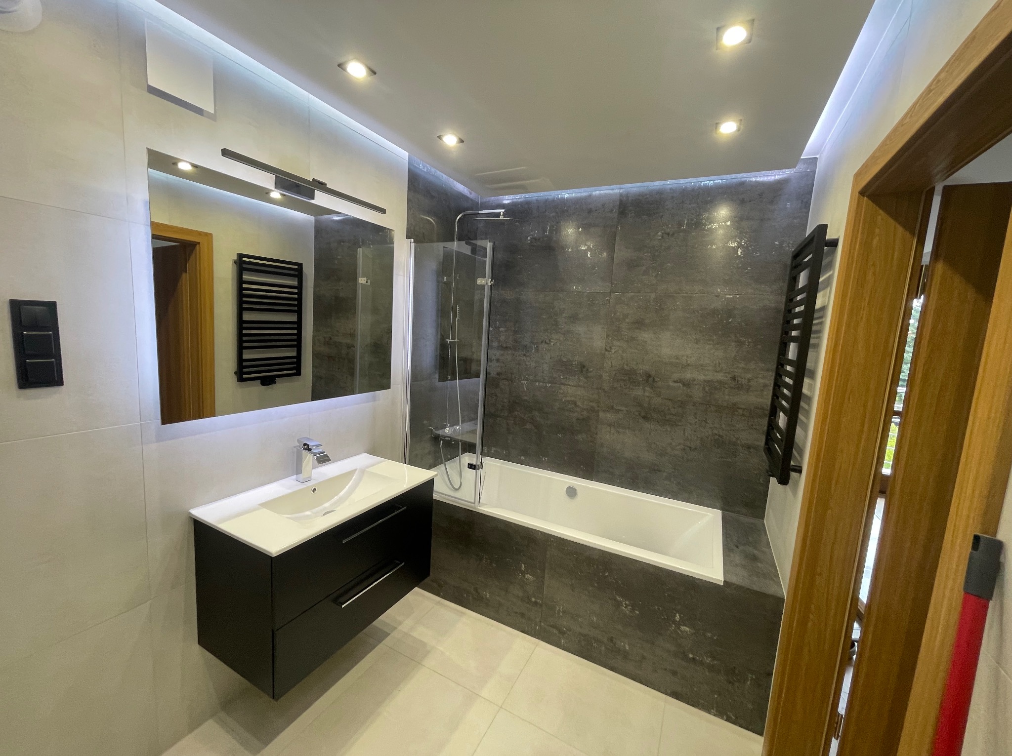 Nowoczesna łazienka z szarymi płytkami imitującymi kamień, białą wanną z parawanem, czarną szafką pod umywalką i dużym lustrem z oświetleniem LED.