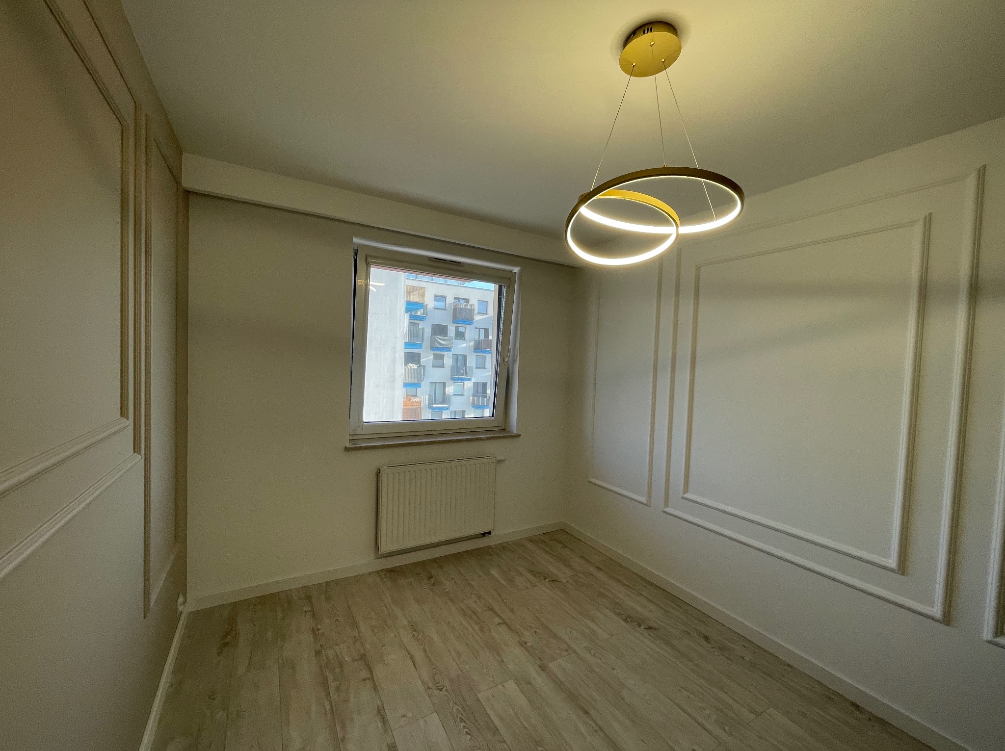 Pusty pokój z panelami ściennymi w stylu klasycznym, parkietem w odcieniach beżu i nowoczesną lampą sufitową z dwoma okręgami LED.