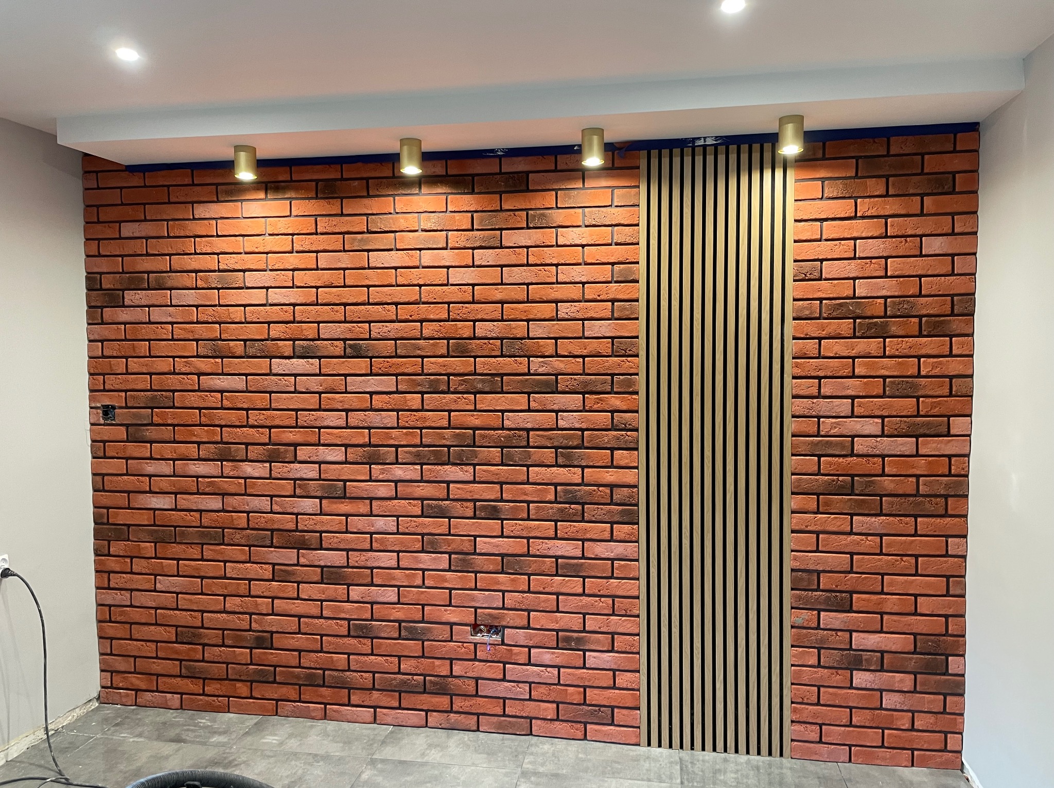 Ściana z cegły klinkierowej z pionowym panelem dekoracyjnym z drewnianych listew, oświetlona punktowymi lampami sufitowymi.