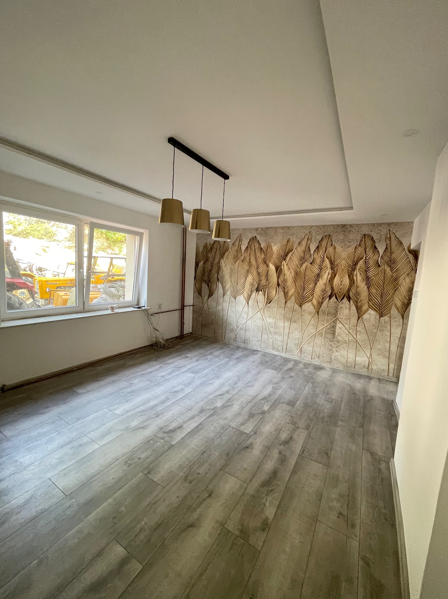 Pusty pokój po remoncie z panelami podłogowymi imitującymi drewno, tapetą w złote liście na jednej ścianie i trzema złotymi lampami wiszącymi.