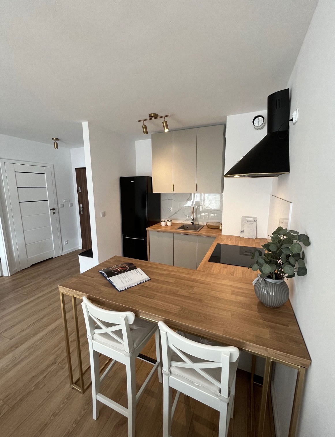 Jasna kuchnia z drewnianym blatem, białymi krzesłami barowymi i czarną lodówką, oświetlona złotymi reflektorami, uzupełniona o książkę i roślinę doniczkową na blacie.