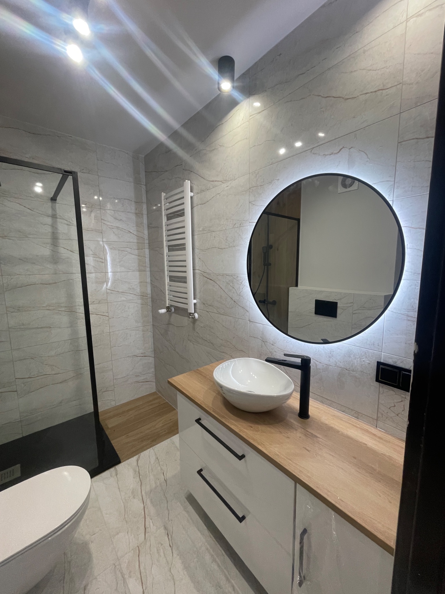 Nowoczesna łazienka z kabiną prysznicową typu walk-in, umywalką nablatową na drewnianym blacie i okrągłym lustrem z podświetleniem LED, wykończona płytkami imitującymi marmur.