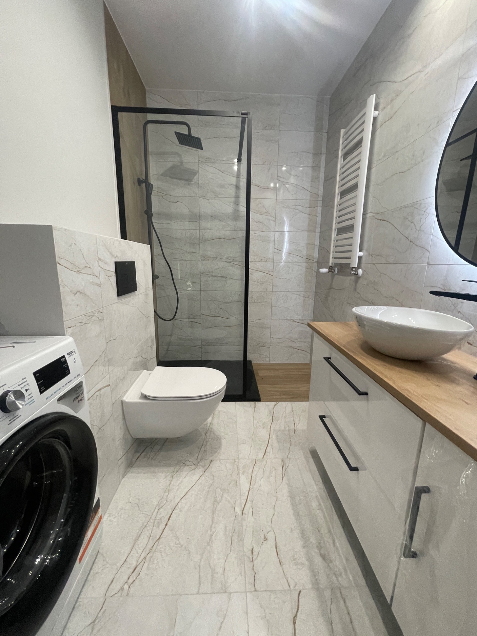 Nowoczesna łazienka wykończona w jasnych marmurowych płytkach z akcentami czerni: kabina prysznicowa typu walk-in z czarną ramą, podwieszana toaleta, umywalka nablatowa na szafce z drewnianym...