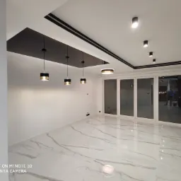 Nowoczesne wnętrze z białymi ścianami, marmurową podłogą, czarnymi akcentami na suficie i wiszącymi lampami z elementami w kolorze złota, widok na przesuwne drzwi balkonowe.