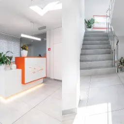 Jasne wnętrze kliniki z recepcją w kolorze białym i pomarańczowym, roślinami doniczkowymi i schodami na piętro.