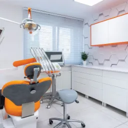 Nowoczesny gabinet stomatologiczny z pomarańczowymi akcentami, widok na fotel dentystyczny, unit, lampę zabiegową i szafki z umywalką na tle heksagonalnych płytek.