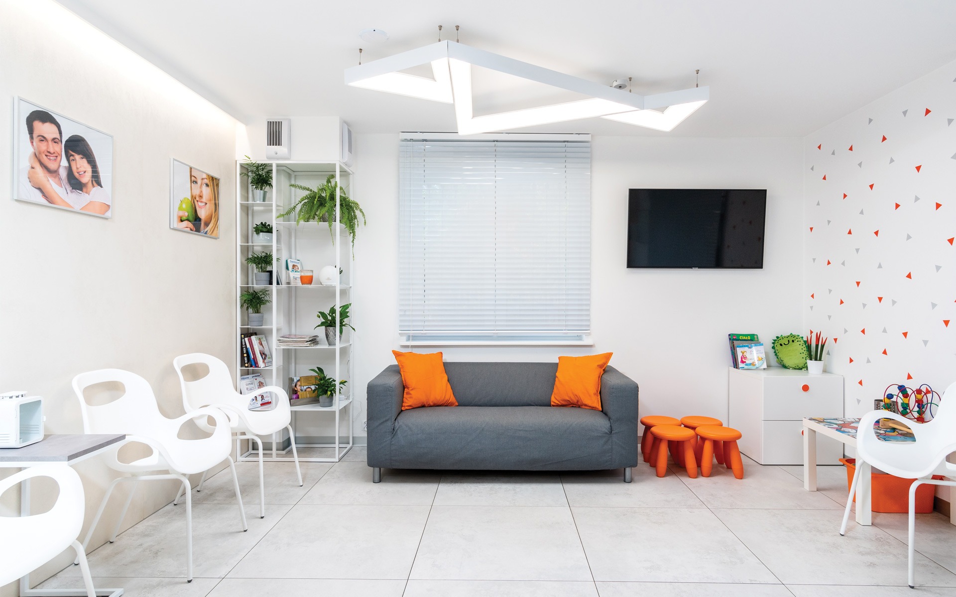 Jasna poczekalnia z szarą sofą z pomarańczowymi poduszkami, białymi krzesłami, regałem z roślinami i dekoracyjnymi naklejkami trójkątów na ścianie.