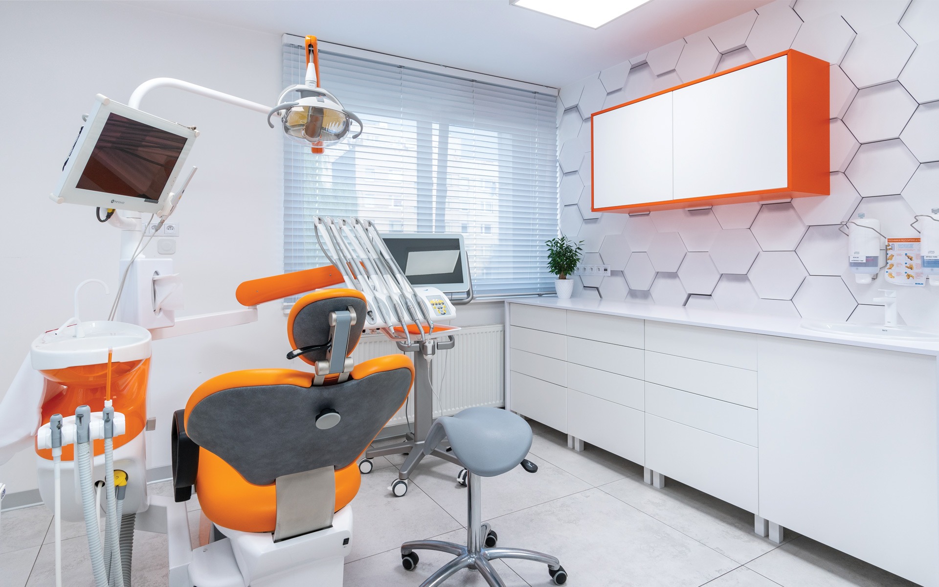 Nowoczesny gabinet stomatologiczny z pomarańczowymi akcentami, widok na fotel dentystyczny, unit, lampę zabiegową i szafki z umywalką na tle heksagonalnych płytek.