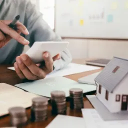 Osoba używa kalkulatora nad stosem monet, dokumentami i modelem domu na biurku, w tle widoczny wykres.
