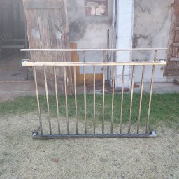 MGweld - Surowa, metalowa konstrukcja balustrady z pionowymi prętami, ustawiona na trawie przed warsztatem z otwartymi drewnianymi wrotami i ścianą z oknem.