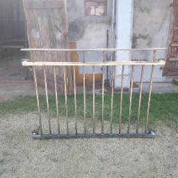 Surowa, metalowa konstrukcja balustrady z pionowymi prętami, ustawiona na trawie przed warsztatem z otwartymi drewnianymi wrotami i ścianą z oknem.