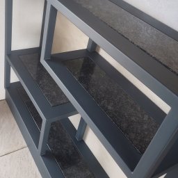 MGweld - Wielopoziomowy metalowy regał w kolorze grafitowym z blatami imitującymi ciemny granit, umieszczony przy jasnej ścianie.