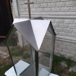 MGweld - Nierdzewna kapliczka z krzyżem na szczycie, szklane ścianki, tło z ceglanego muru i drewnianych drzwi.