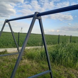 MGweld - Metalowa konstrukcja huśtawki ogrodowej w kolorze grafitowym, ustawiona na trawniku z widokiem na pole uprawne i ogrodzenie panelowe w tle.