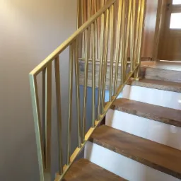 Złota metalowa balustrada schodowa z pionowymi szczeblami, zamontowana na drewnianych stopniach w jasnym wnętrzu.