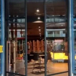 Otwarta, dwuskrzydłowa brama z czarną, metalową ramą i przezroczystym wypełnieniem szklanym, prowadząca do wnętrza budynku. Widoczne elementy konstrukcyjne i wyposażenie wnętrza w tle.