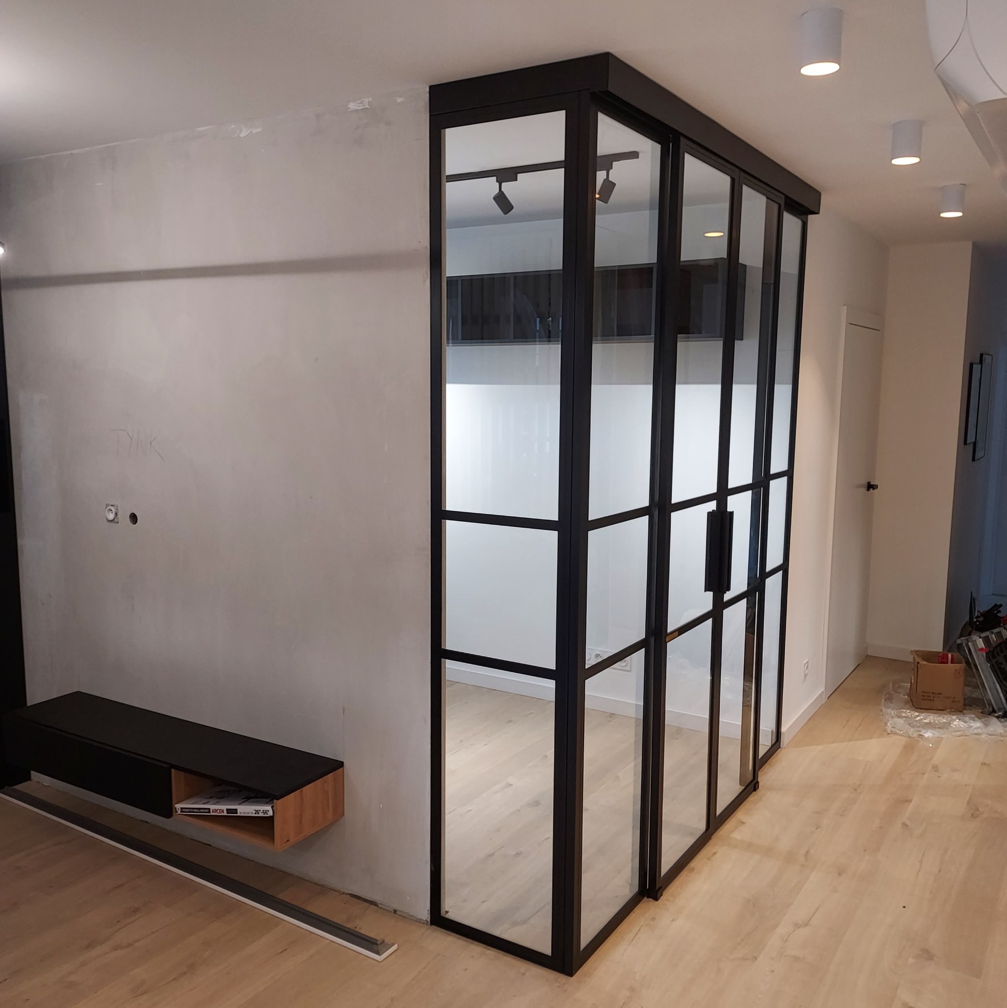 Nowoczesna szklana ścianka działowa z czarnymi profilami stalowymi w jasnym wnętrzu, widoczna niedokończona ściana z napisem 'TYNK' i minimalistyczna szafka RTV.