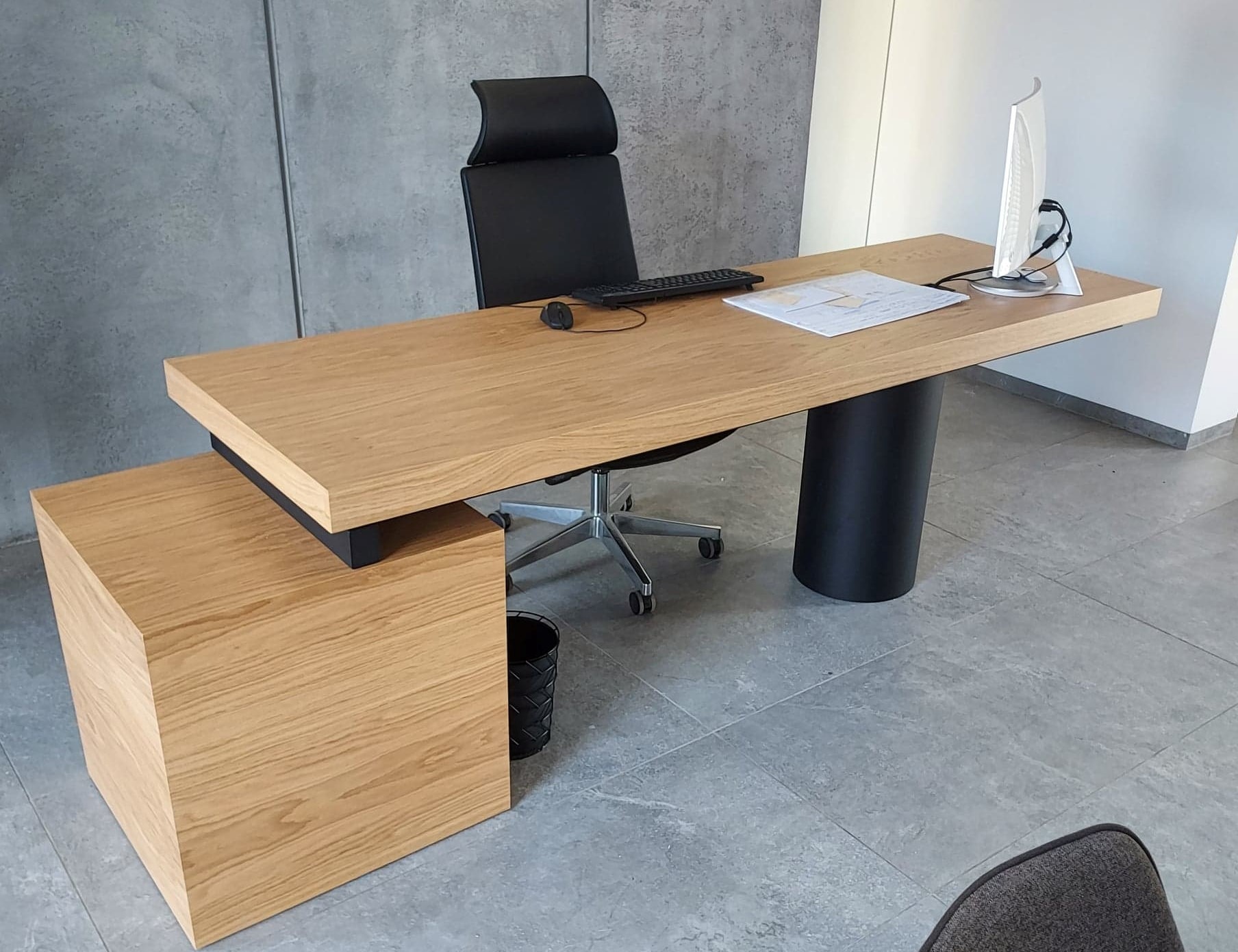 Nowoczesne biurko z blatem z jasnego drewna, wsparte na czarnej, cylindrycznej nodze, z dostawką w formie szafki; na blacie klawiatura, mysz i dokumenty, w tle szary beton architektoniczny.