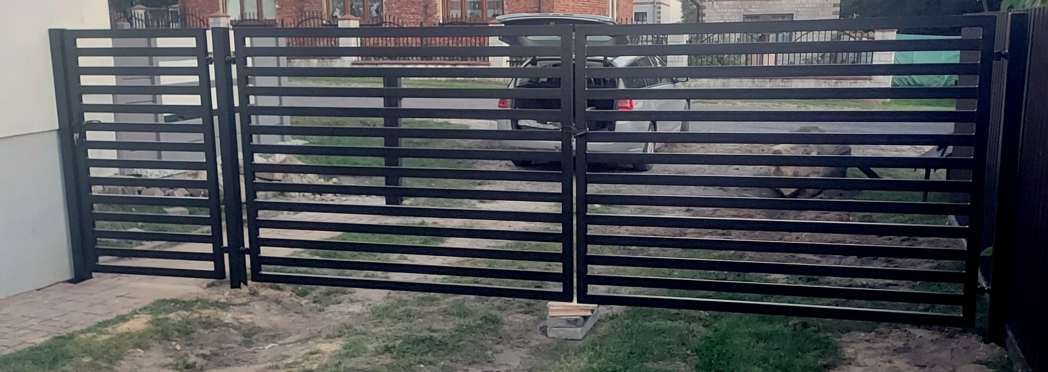 Nowoczesna, pozioma brama wjazdowa i furtka z czarnych, płaskich profili, widok z zewnątrz na posesję z zaparkowanym samochodem.