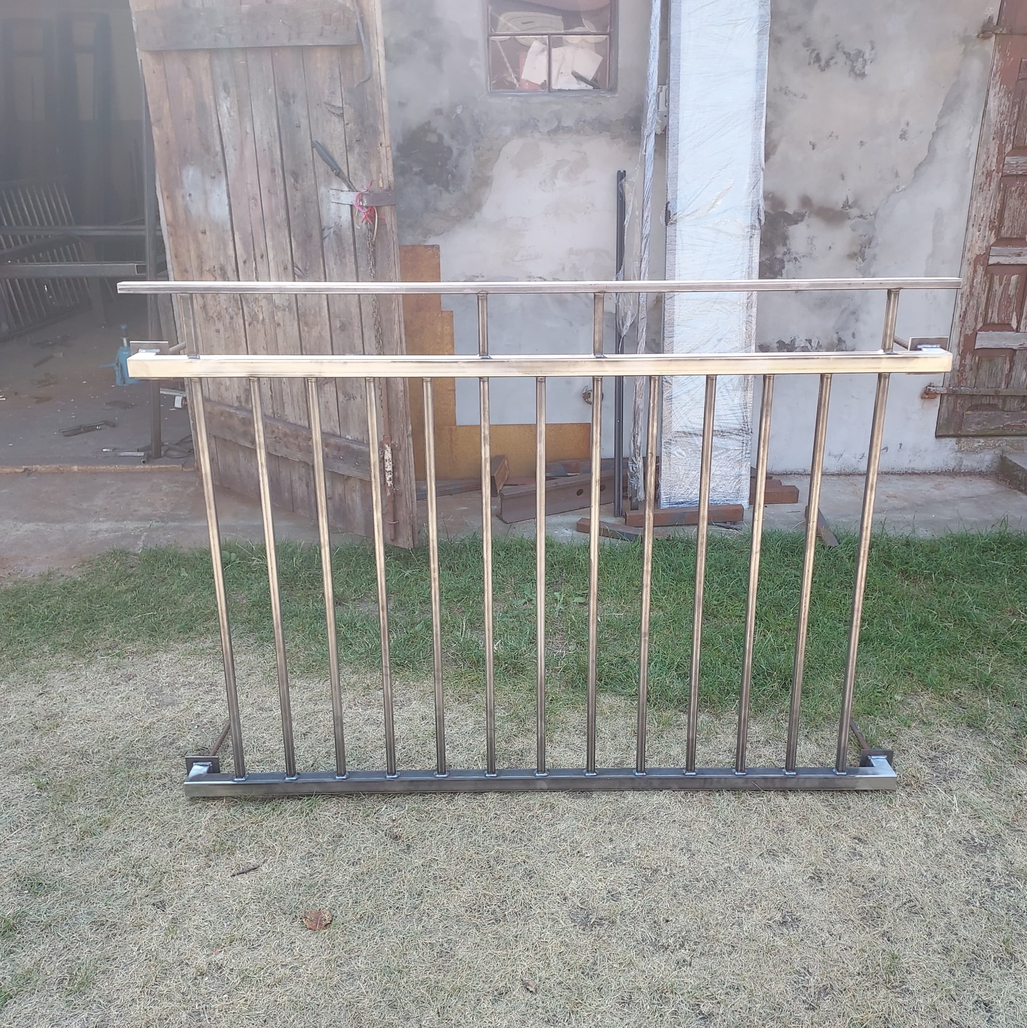 Surowa, metalowa konstrukcja balustrady z pionowymi prętami, ustawiona na trawie przed warsztatem z otwartymi drewnianymi wrotami i ścianą z oknem.